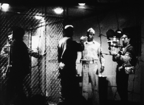 A black-and-white photo depicting a behind the scenes view of a movie in production. A man holds the camera on the right - a cage is on the left, and two men speak in the center of the image, one facing the camera and one facing away.
