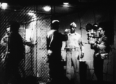 A black-and-white photo depicting a behind the scenes view of a movie in production. A man holds the camera on the right - a cage is on the left, and two men speak in the center of the image, one facing the camera and one facing away.