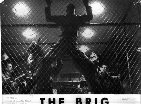 A man clinging to a chain-link fence in front of several other men looking up at him. Underneath the image, partially cut-off text reads, "THE BRIG."