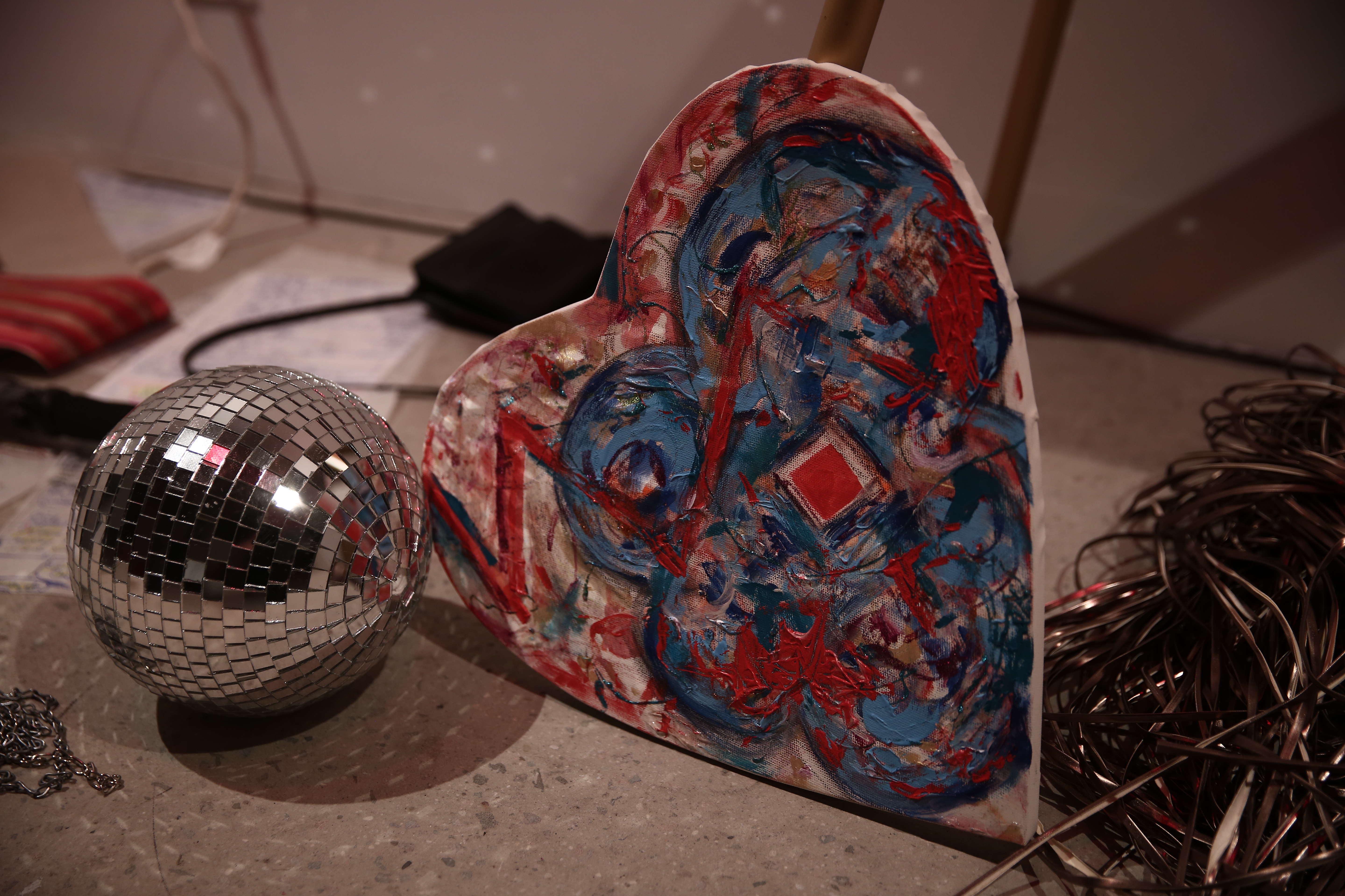 Various objects sit on a concrete floor, including a miniature disco ball, a painted heart-shaped canvas, and a tangle of cords.