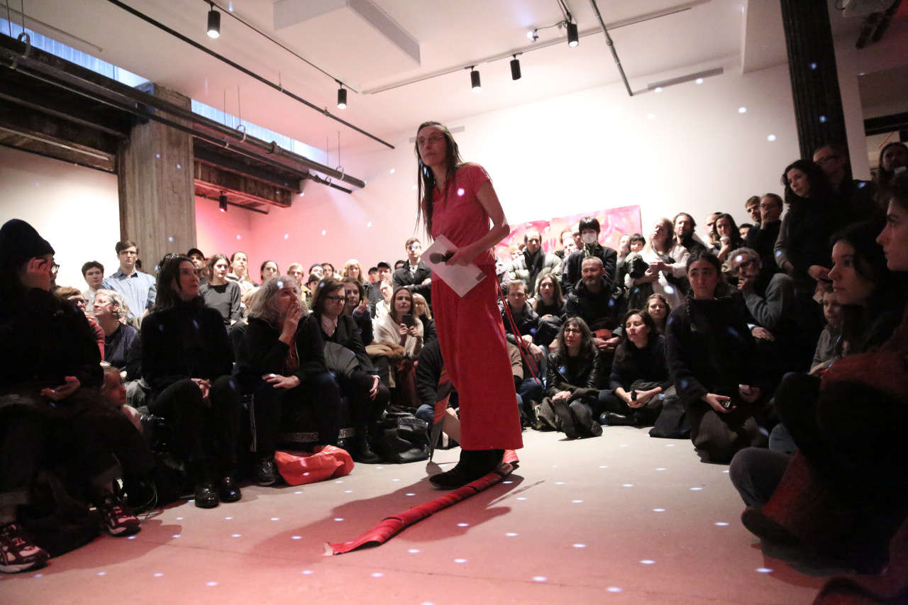 A person dressed in all pink, holding a microphone and piece of paper, stands surrounded by a crowd of audience members arranged in a circle. The room is lit with a slightly pink light and light spots are reflected at even intervals.