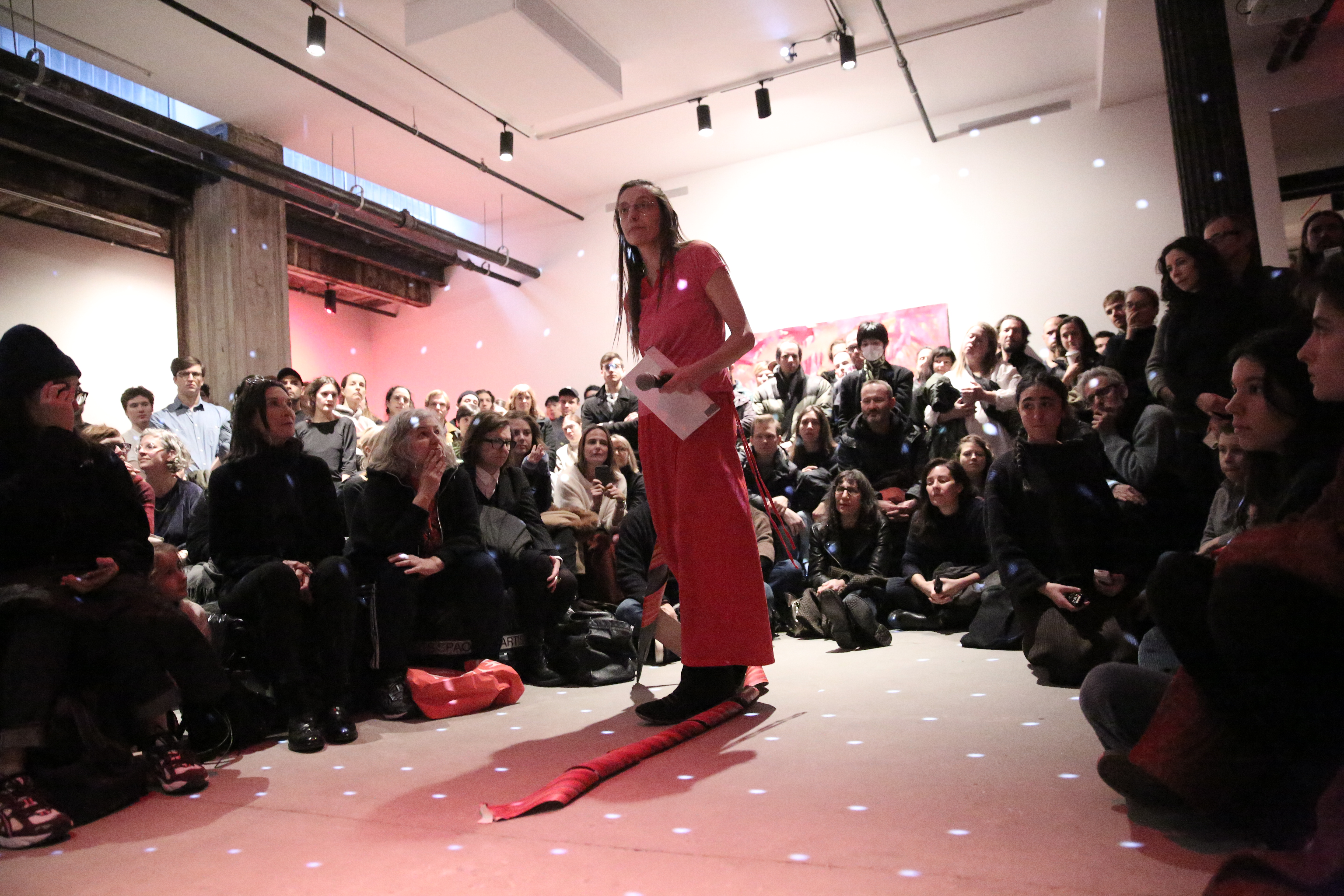 A person dressed in all pink, holding a microphone and piece of paper, stands surrounded by a crowd of audience members arranged in a circle. The room is lit with a slightly pink light and light spots are reflected at even intervals.
