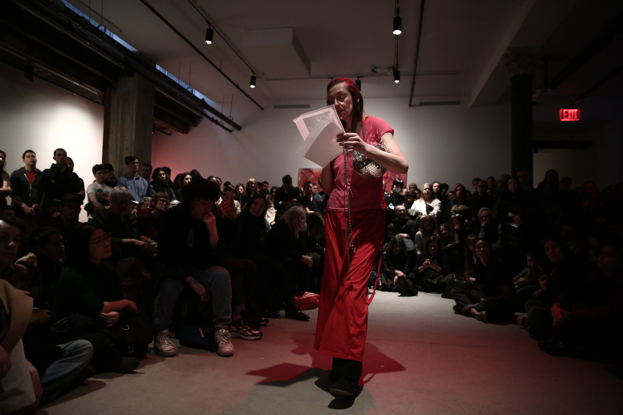 A person dressed in all pink reads from a sheet of paper into a microphone. She holds a small disco ball in her arm. She is surrounded by rapt audience members arranged in a semi-circle around her.