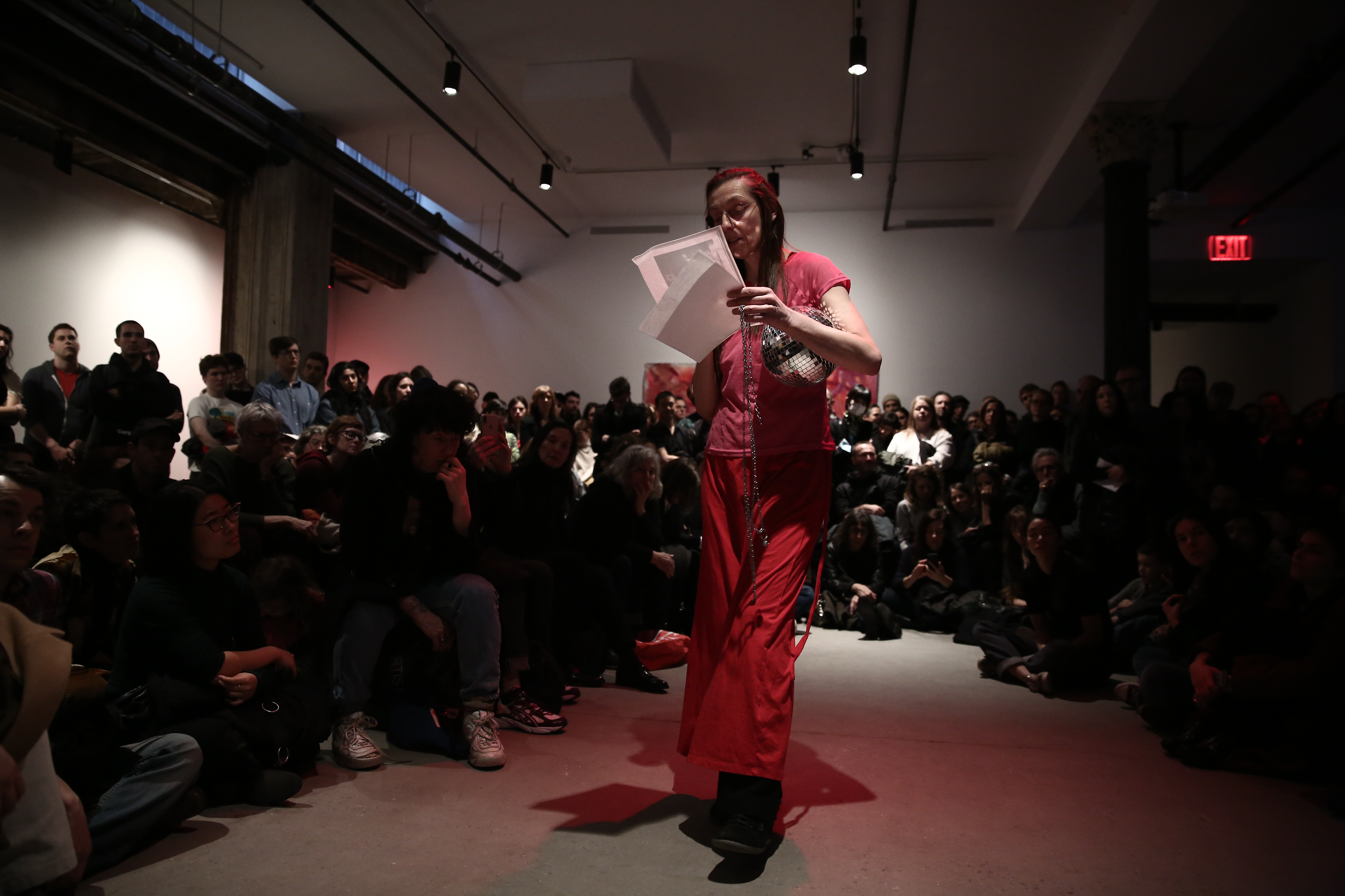A person dressed in all pink reads from a sheet of paper into a microphone. She holds a small disco ball in her arm. She is surrounded by rapt audience members arranged in a semi-circle around her.