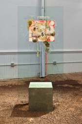 An installation view of a brightly colored painting lodged into a rectangular sheet of glass which is made to stand upright by being wedged into a block of concrete which sits on a floor scattered with wood chips.