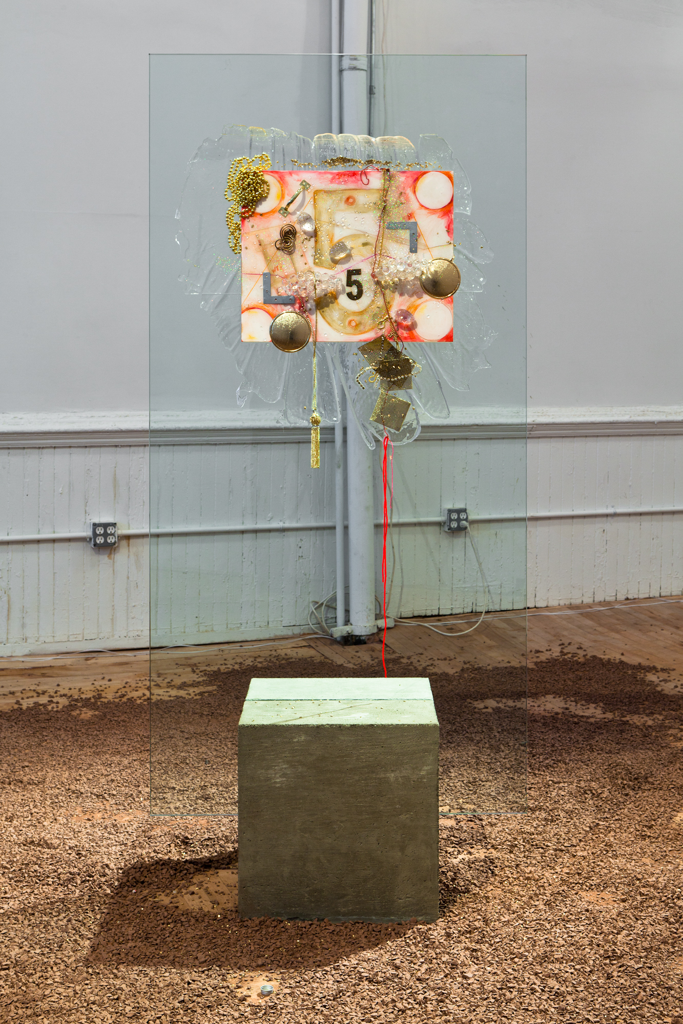 An installation view of a brightly colored painting lodged into a rectangular sheet of glass which is made to stand upright by being wedged into a block of concrete which sits on a floor scattered with wood chips.