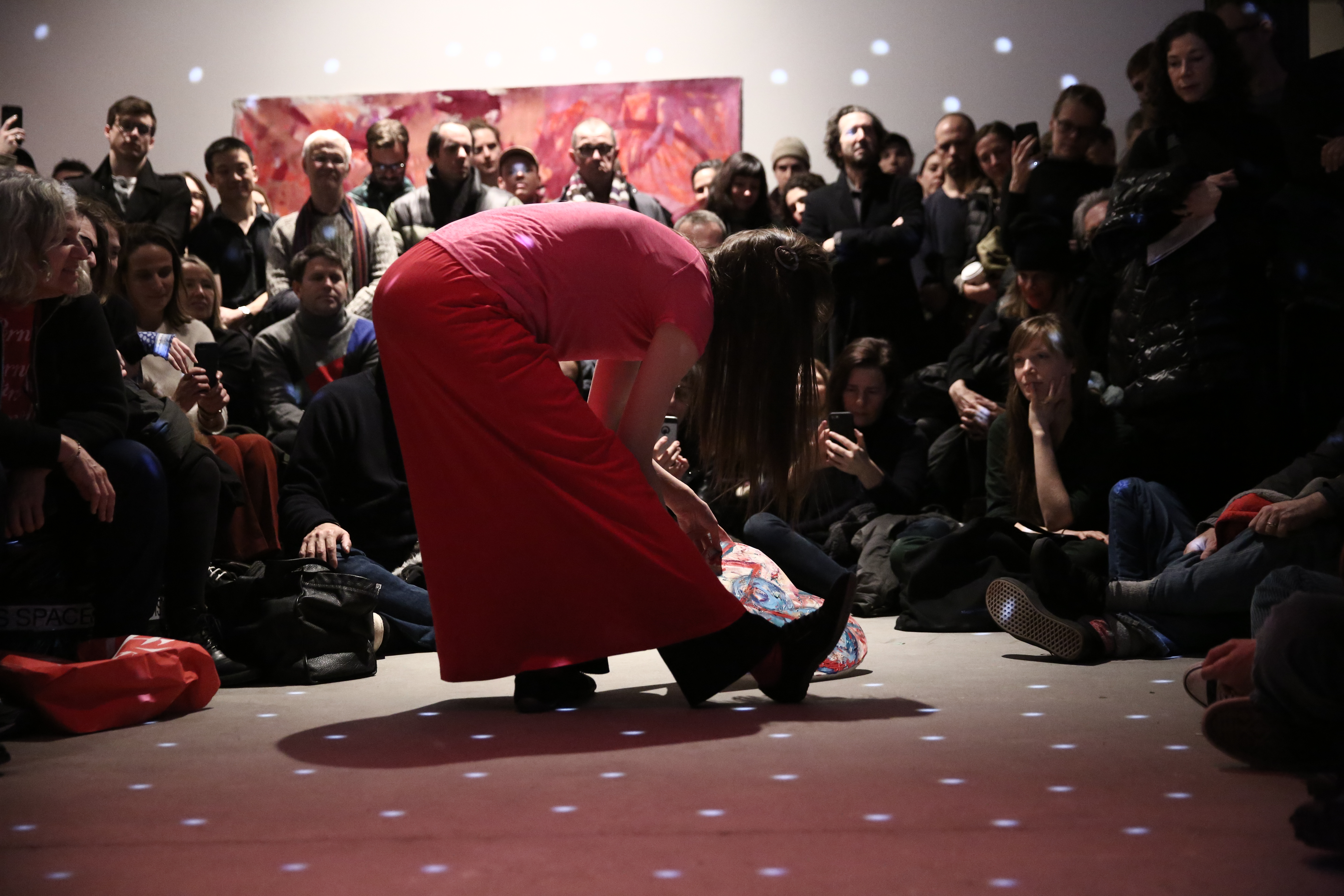 A person dressed in all pink bends at the waist over her extended leg, over a heart-shaped canvas. She is surrounded by a crowd of audience members. There are light spots are reflected at even intervals over the performer and the crowd.