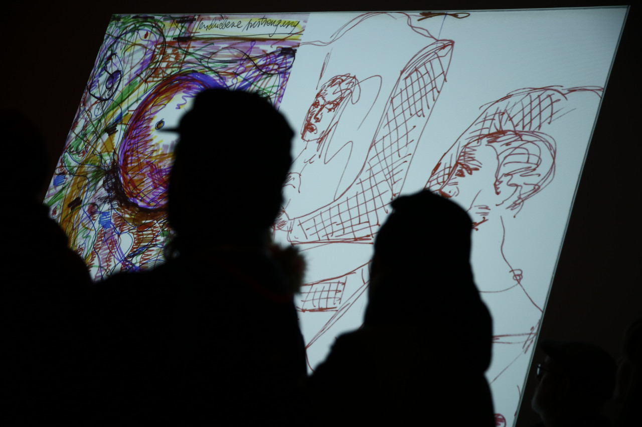 A colorful drawing with sketches of two people is projected onto a dark wall. The image is partially obscured by two silhouetted figures.