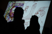 A colorful drawing with sketches of two people is projected onto a dark wall. The image is partially obscured by two silhouetted figures.