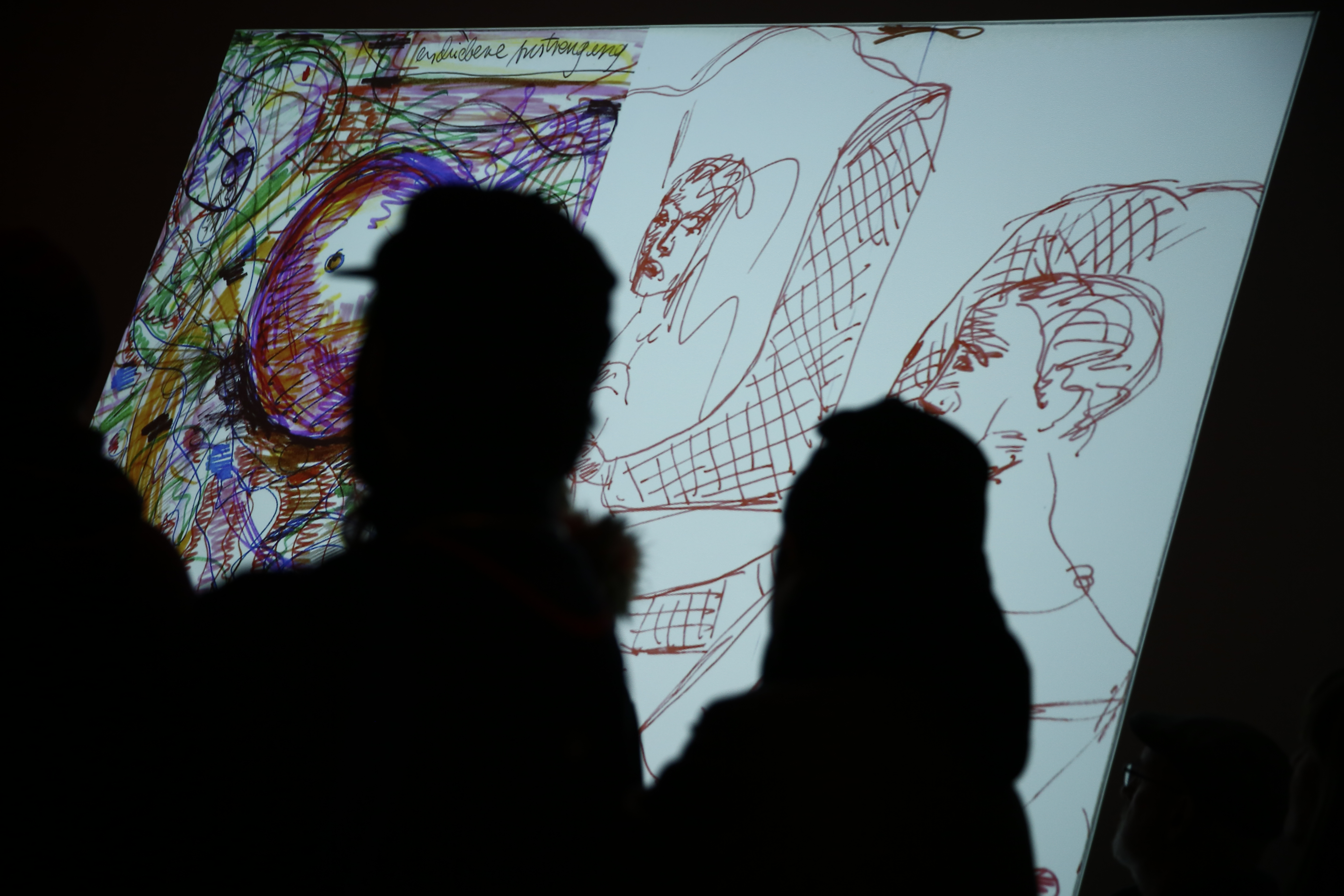 A colorful drawing with sketches of two people is projected onto a dark wall. The image is partially obscured by two silhouetted figures.