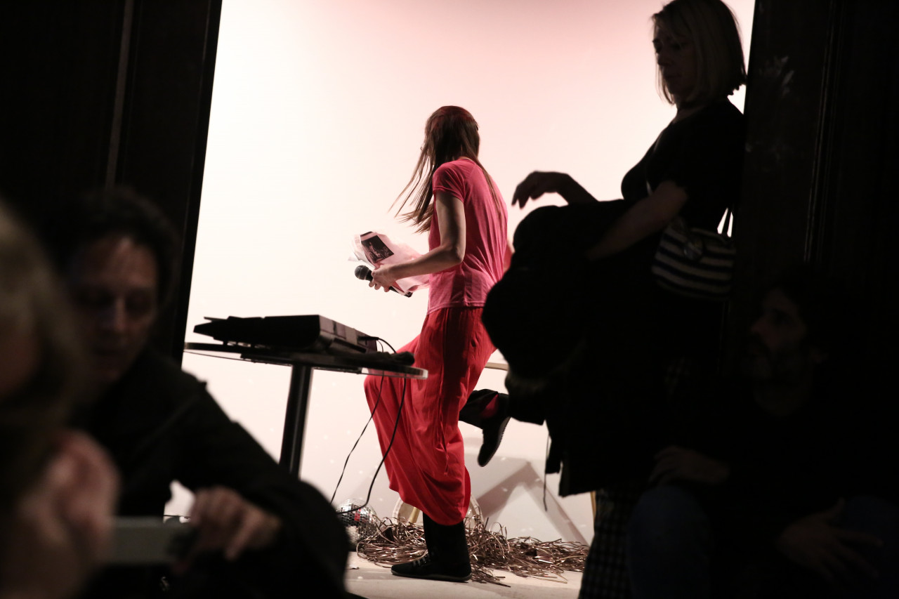 A person dressed in all pink steps over a mass of wires. She is illuminated from behind by a bright, white wall. In the foreground, dimly lit figures face varying directions.