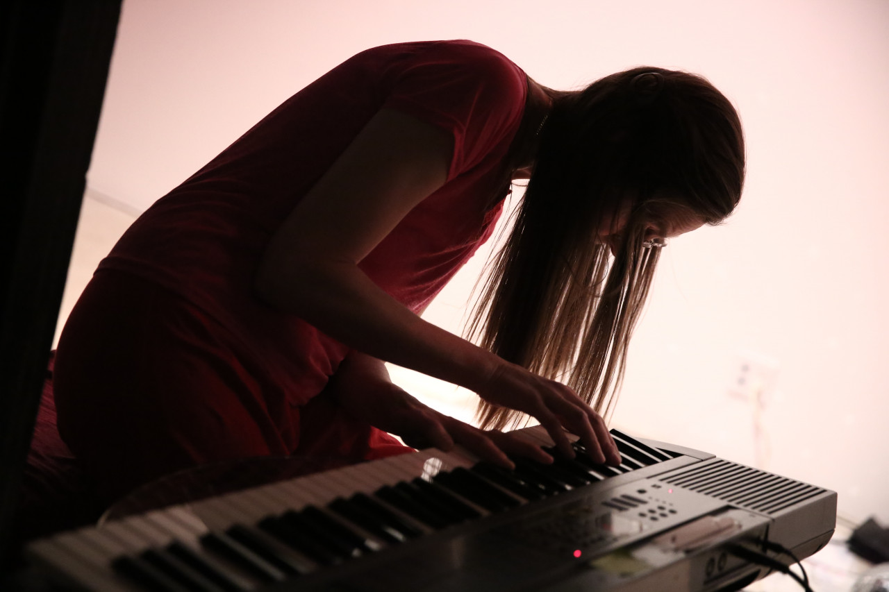 A person bends over an electric piano, her hair falling over her face. She is illuminated from behind by a brightly lit wall.