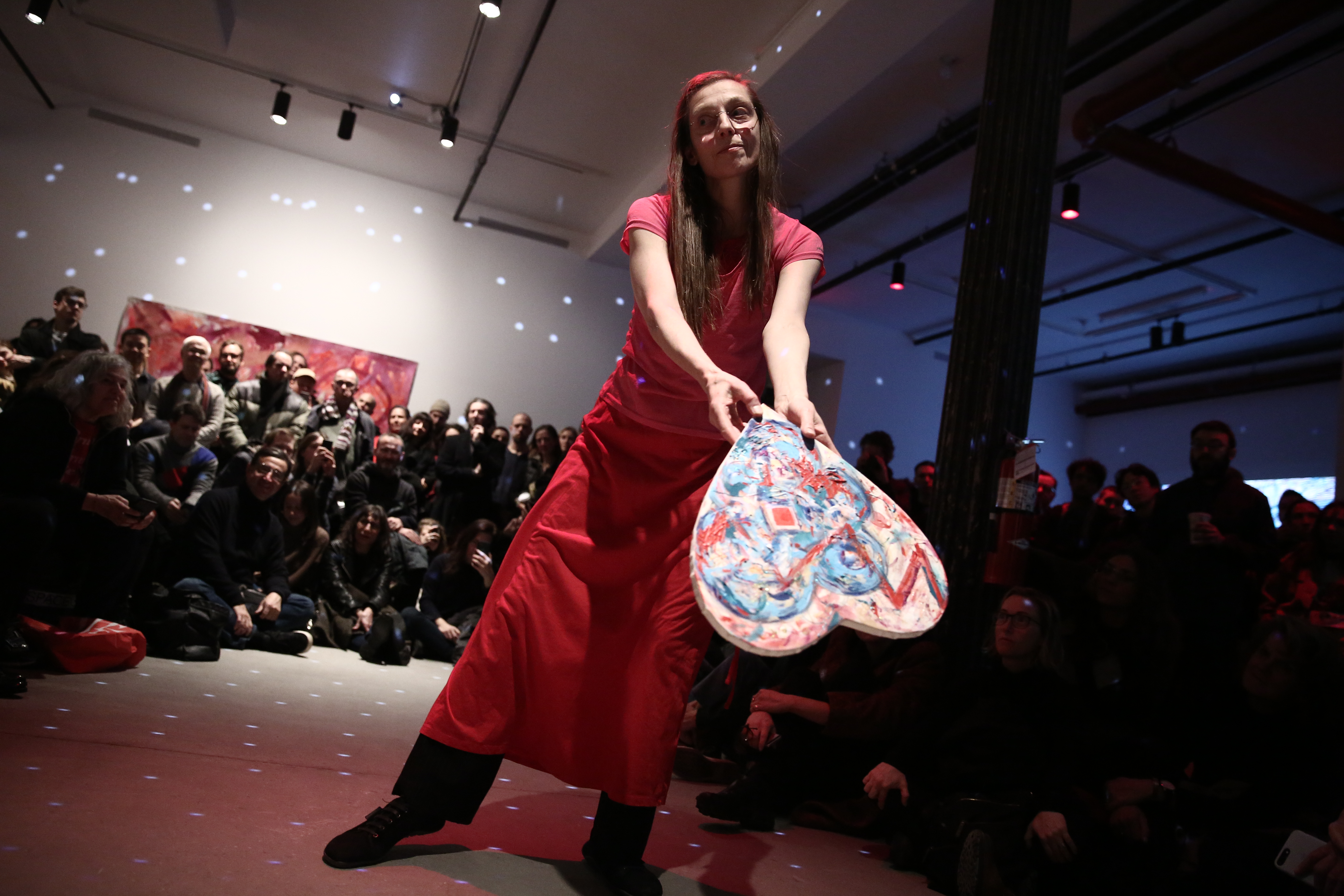A person dressed in all pink extends a heart-shaped canvas in front of her, while her eyes look away. She is surrounded by rapt audience members arranged in a semi-circle around her. There are light spots reflected at even intervals patterning the room.