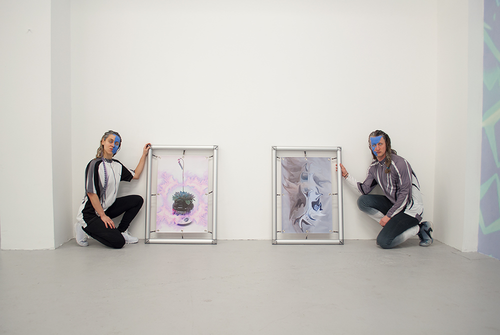 Two figures crouch next to two abstract artworks standing on the floor of a gallery space. Both people have dyed hair and half-blue facepaint.