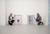 Two figures crouch next to two abstract artworks standing on the floor of a gallery space. Both people have dyed hair and half-blue facepaint.