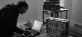 A man looking at a laptop, with a modular synth and mixer next to the laptop.