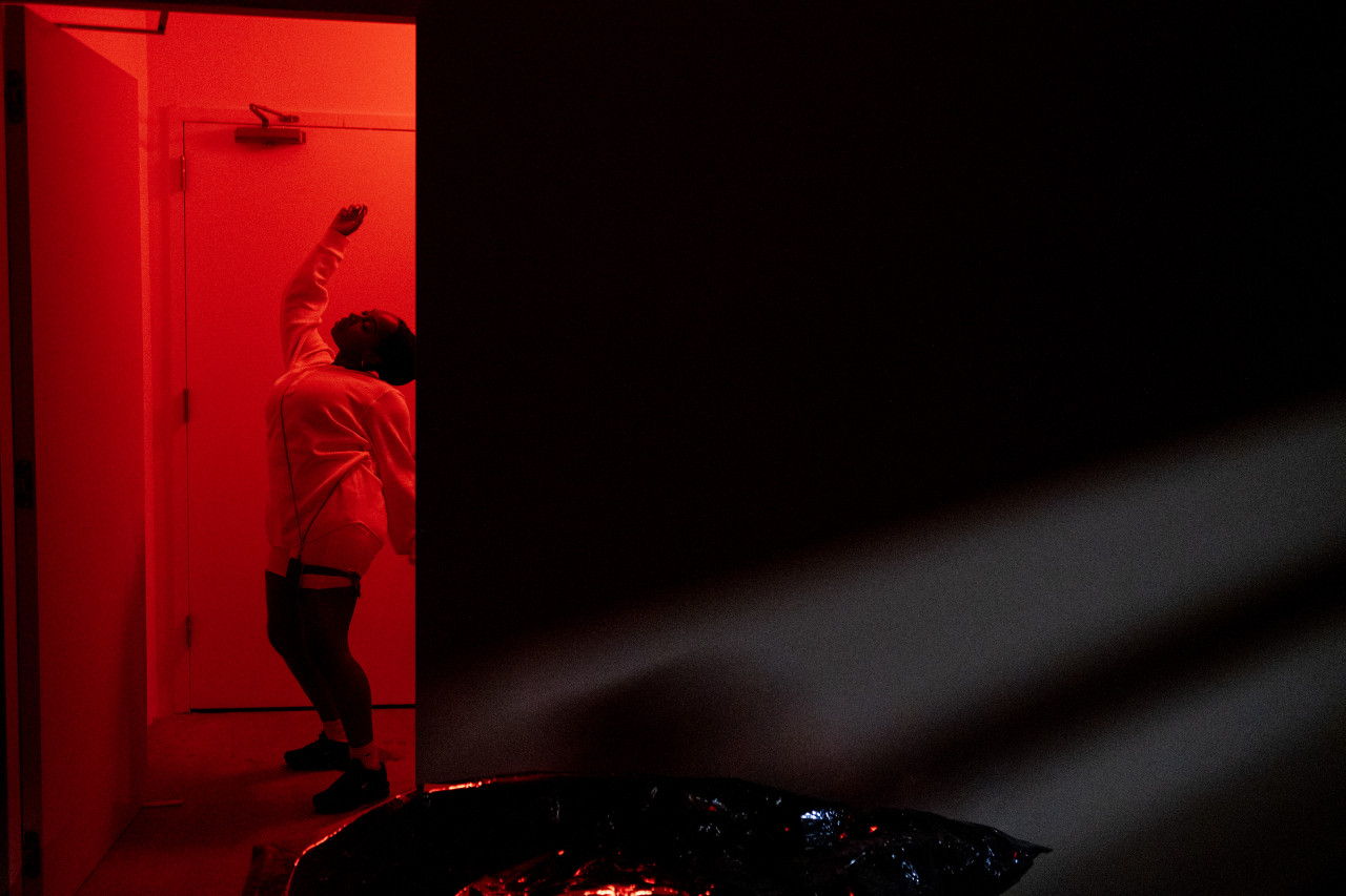 Color photograph of a figure dressed in all white, dancing in a corridor bathed in red light.