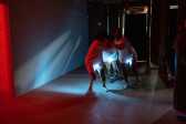 Color photograph of three figures dressed in white, embracing each other as they run across an empty basement.