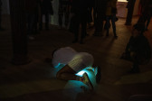 Color photograph of a performer dressed in white, kneeling on all fours. A bright light from their pelvic area shines onto the floor, as viewers stand around the figure.