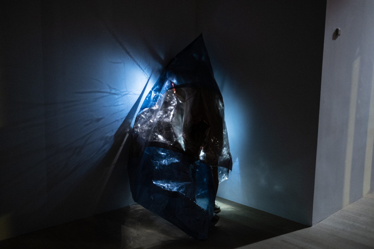 Color photograph of a figure wearing a conical sheath of reflective one-way mirror film. They stand in the corner of a dark room, shining a bright light against the wall behind them. Their silhouette can be vaguely seen under the mirror film.