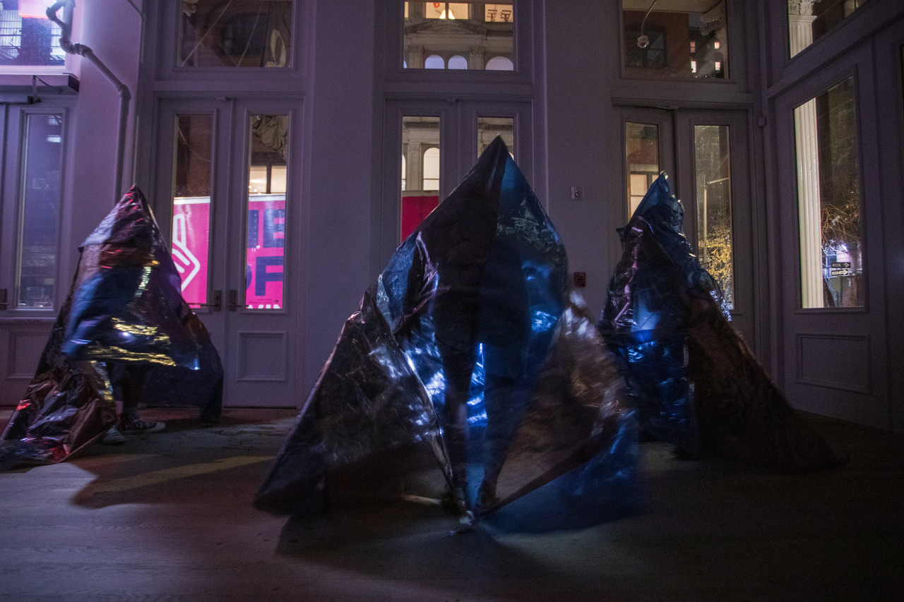 Color photograph of three figures, wearing conical costumes made of reflective one-way mirror film, standing at the doorways of a gallery space.