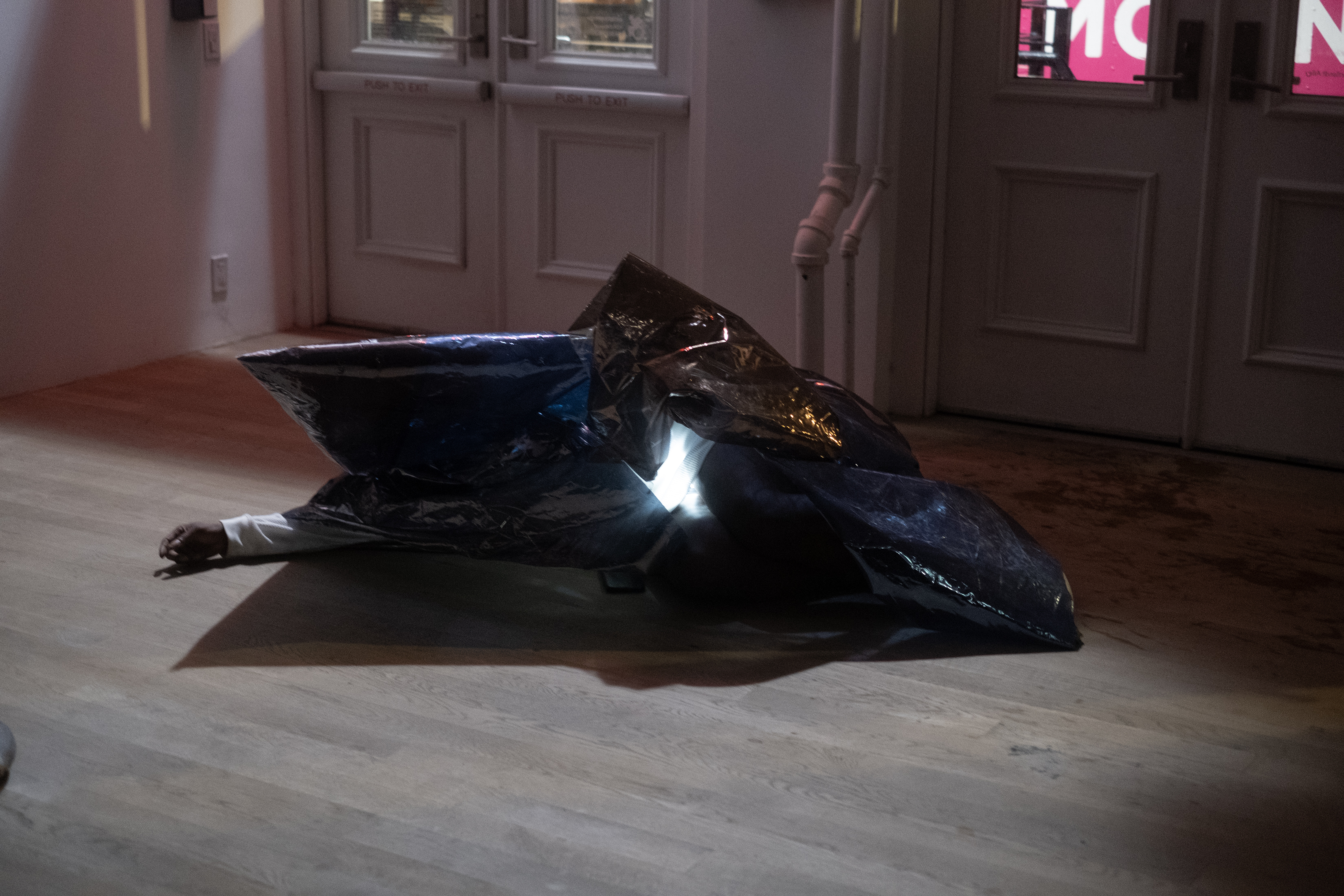 Color photograph of a figure wearing a conical sheath of reflective one-way mirror film that covers most of their body. They lay on the floor and spread one arm out from under the mirror film. A bright light shines from under the film.