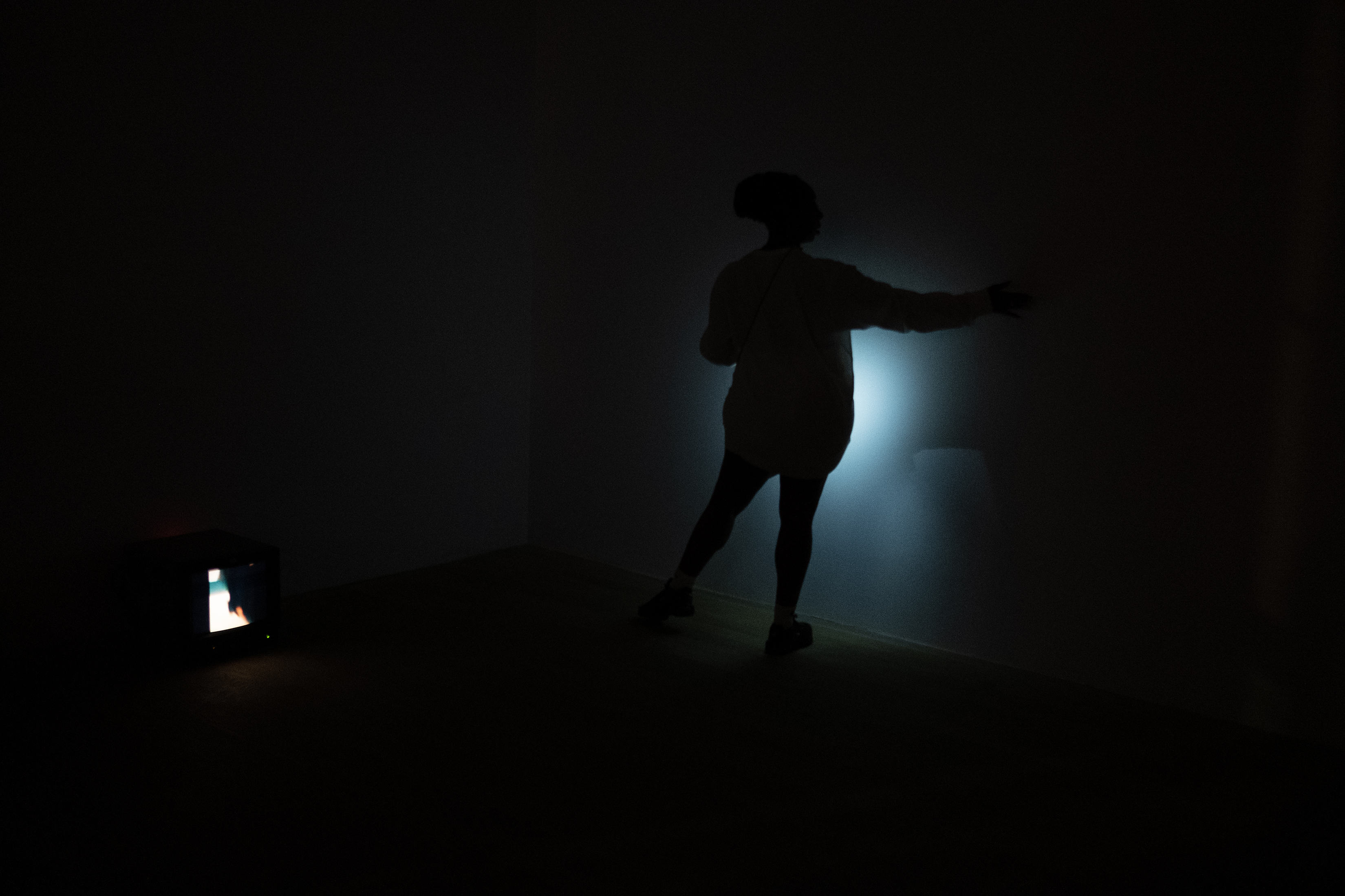 Color photograph of a figure in a completely dark room, facing one wall and shining a light from their body against the wall.