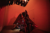 Color photograph of a corner space in a gallery, below a staircase, that is bathed in red light. At the bottom of the stairs are a group of performers wearing conical costumes made of reflective one-way mirror film.