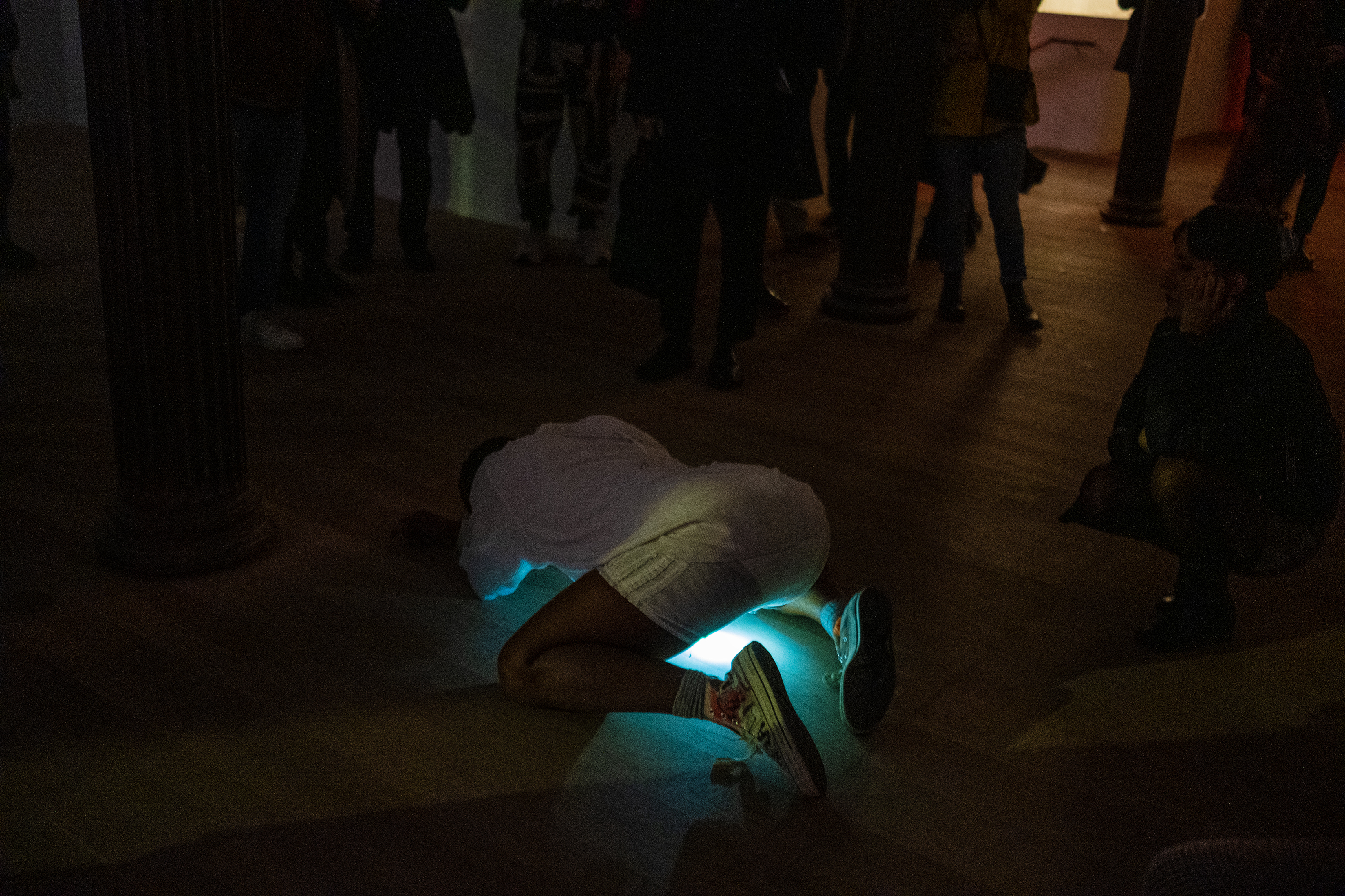 Color photograph of a performer dressed in white, kneeling on all fours. A bright light from their pelvic area shines onto the floor, as viewers stand around the figure.