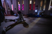 Color photograph of three figures, wearing all white, performing in a dark gallery space. The leftmost figure lays on the floor with their legs spread and hands pushing their chest up. On the right, a figure with a flashlight around their hips stands with their shoulders and head flung back. The third figure contorts their body on the floor in the rightmost corner.