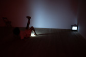 Color photograph of a figure dressed in white, laying on the floor of a dark room. The figure has one leg raised in the air, shining a light from their pelvic area onto the floor and wall across from them. A small TV monitor sits in the corner.