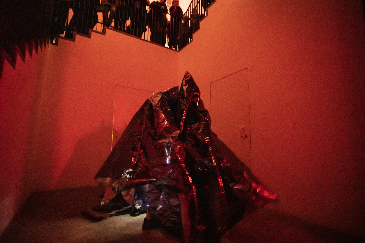Color photograph of a corner space in a gallery, below a staircase, that is bathed in red light. At the bottom of the stairs are a group of performers wearing conical costumes made of reflective one-way mirror film.