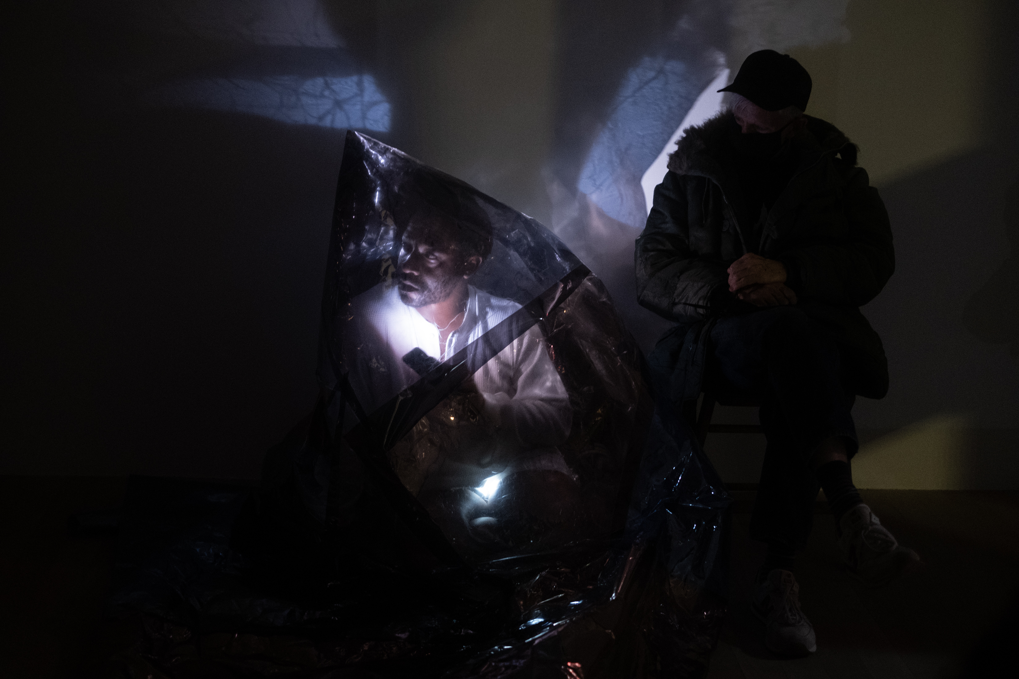 Color photograph of a fgure wearing a conical sheath of reflective one-way mirror film that covers their entire body. They squat down next to a viewer sitting in a wooden chair. The performer shines a bright light against their face, making their face visible from under the mirror film.