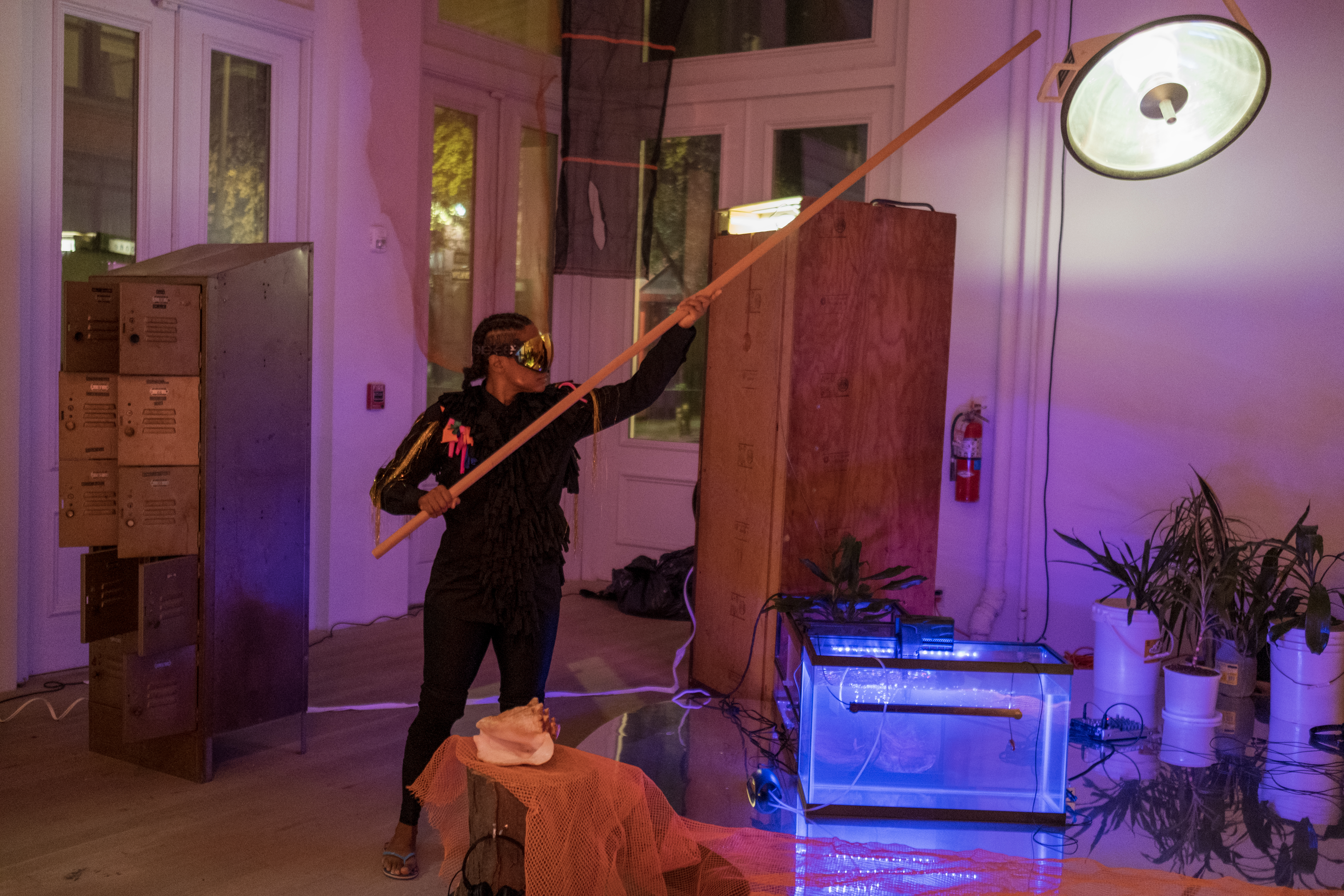 Color photograph of a figure wearing black, holding a long wooden stick over a reflective surface with an orange construction net over it.