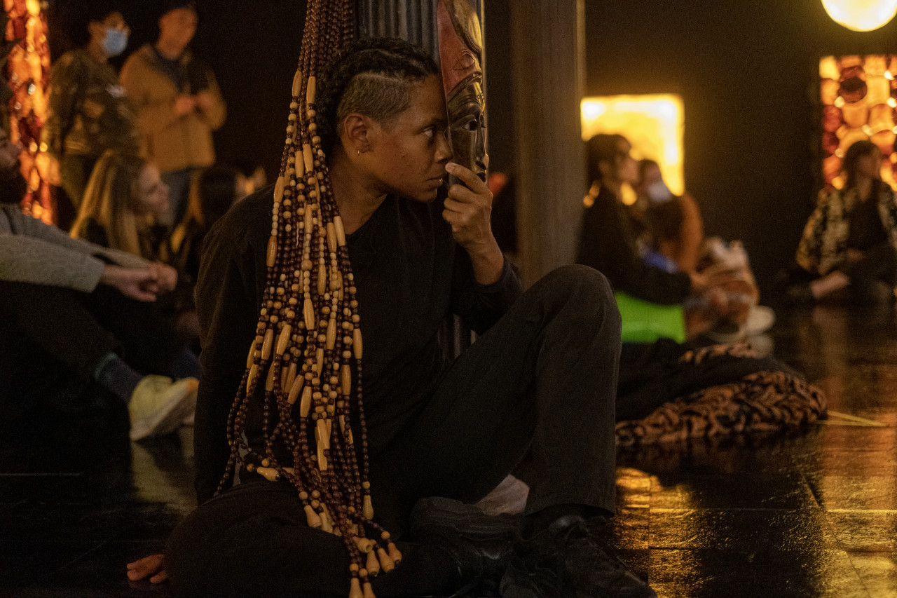 Color photograph of a figure dressed in black performing in front of an audience in the background. The room is dark, with LED light sculptures made from dried SCOBY in the background. The figure sits on the floor, looking off to the right and holding a mask in front of their face.