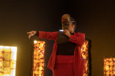 Color photograph of a figure in a red suit performing in a darkly lit room with LED light sculptures in the background. The figure holds their left hand out in a gesture, and with their right hand holds a mask in front of their face.