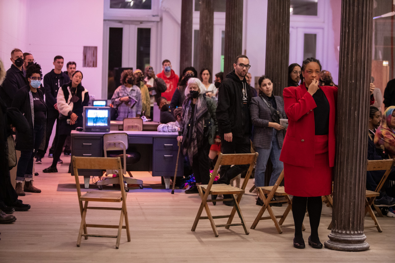 Color photograph of a large group of people standing around a gallery space. Chairs, and tanker desks with TV monitors on them are placed in the space. The audience watches a figure in a red suit that stands next to a column, holding their hand to their mouth.