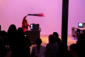 Color photograph of a figure in a red suit preforming in front of a seated audience. The figure sits at a tanker desk with a TV monitor placed on top of it. They wave a red object in the air.