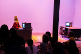Color photograph of a figure in a red suit preforming in front of a seated audience. The figure sits at a tanker desk with a TV monitor placed on top of it. They have papers taped and placed over their face.