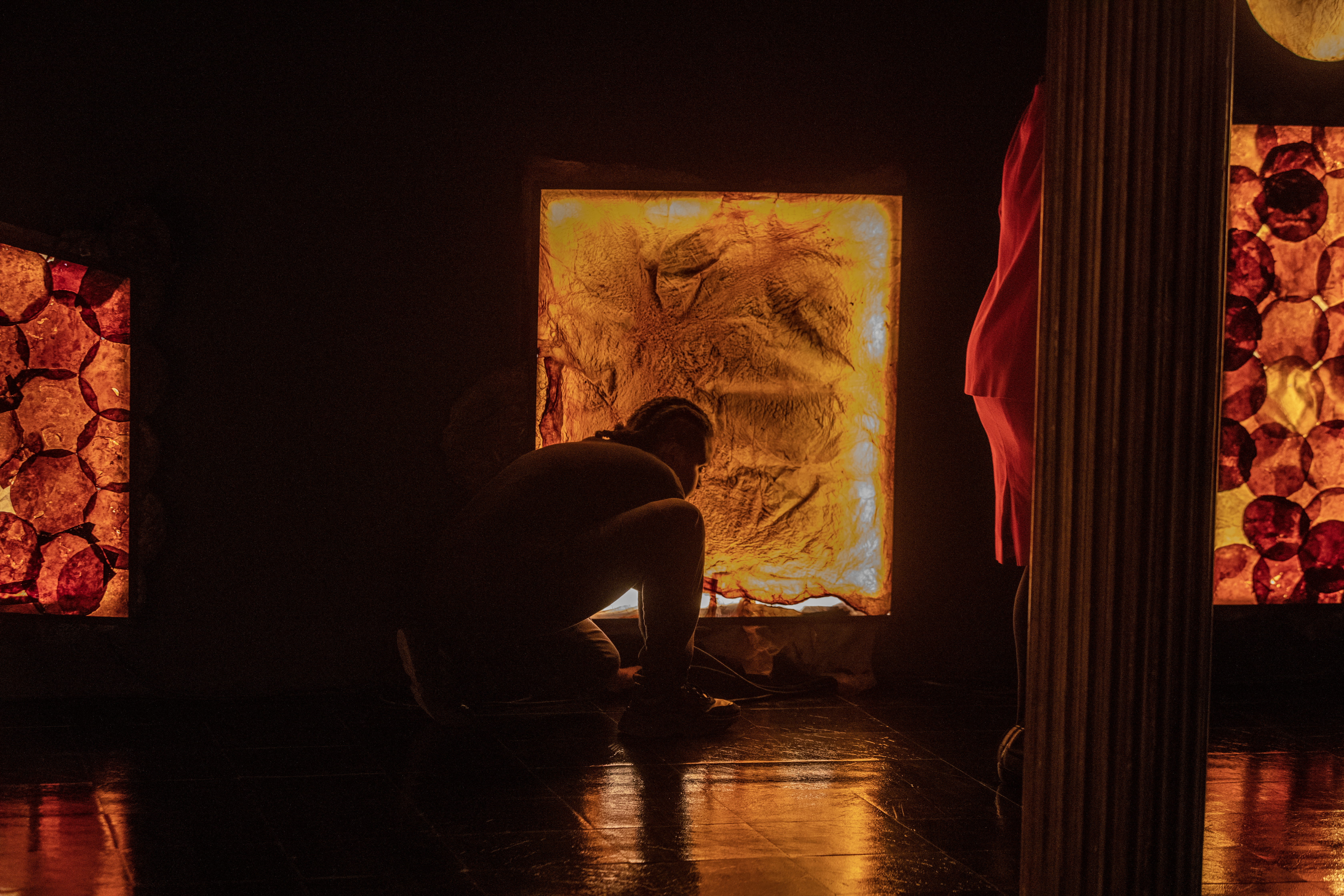 Color photograph of two figures performing in a dark gallery space, lit only by an installation of LED lights covered in dried SCOBY material. One figure wearing black leans down in front of one of the LED lights. Partly hidden behind a column, another figure stands wearing a red suit.