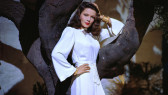 A woman in a white dress with gold bracelets and red lipstick leans against a tree. Her left arms rests on a tree branch, and her right hand is on her hip.