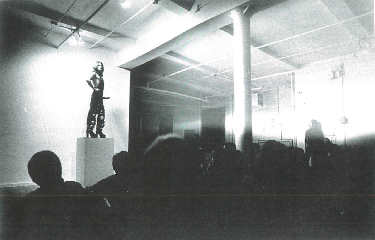 Black and white scan of a photograph of a figure standing on a tall square plinth in a gallery space, in front of an obscured crowd of viewers. The figure poses like a statue with their hands on their hips, wearing a pair of overalls and high, blocky heels, and a phallic strap-on around their waist.