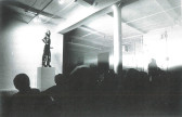 Black and white scan of a photograph of a figure standing on a tall square plinth in a gallery space, in front of an obscured crowd of viewers. The figure poses like a statue with their hands on their hips, wearing a pair of overalls and high, blocky heels, and a phallic strap-on around their waist.