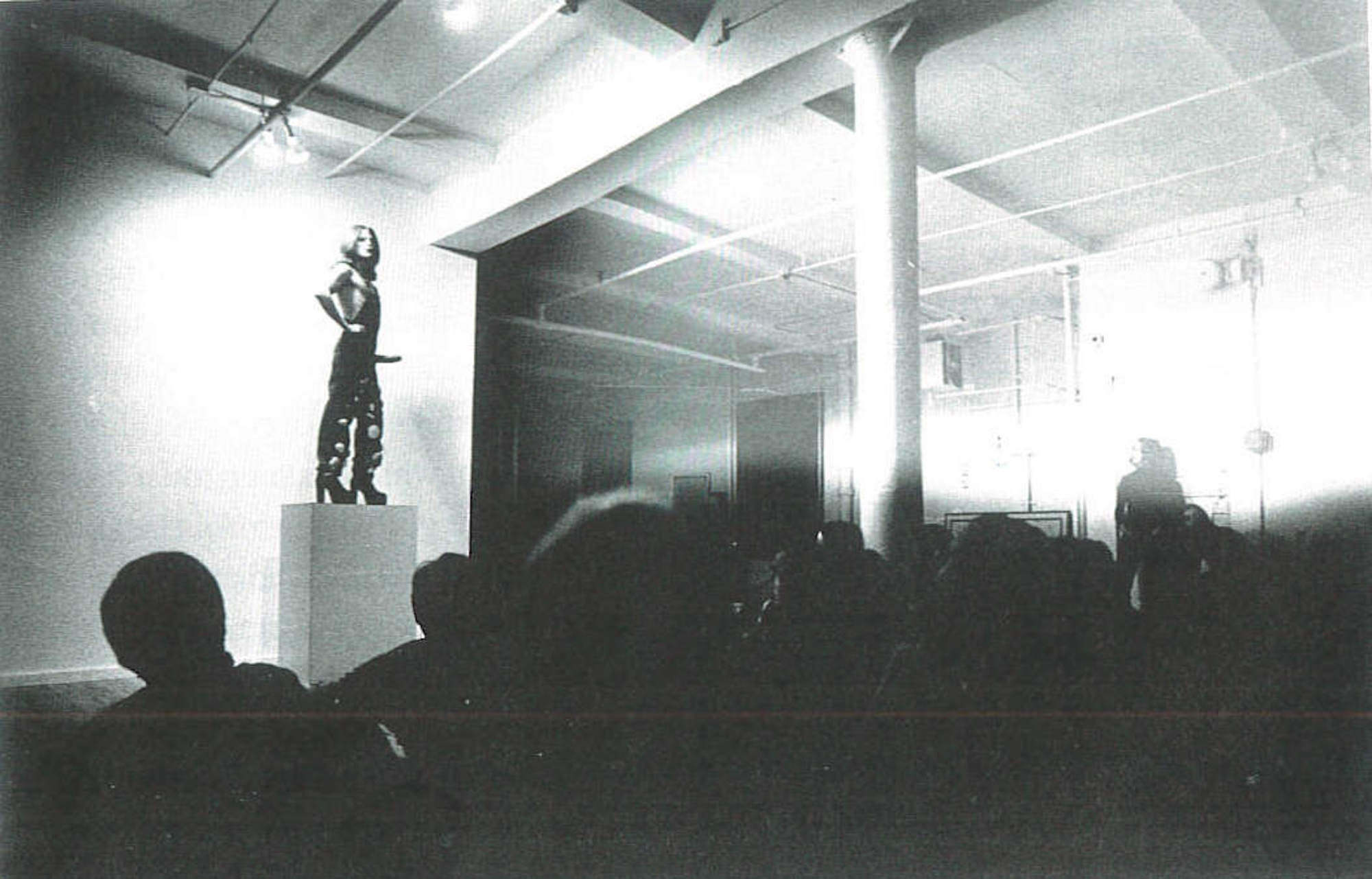 Black and white scan of a photograph of a figure standing on a tall square plinth in a gallery space, in front of an obscured crowd of viewers. The figure poses like a statue with their hands on their hips, wearing a pair of overalls and high, blocky heels, and a phallic strap-on around their waist.