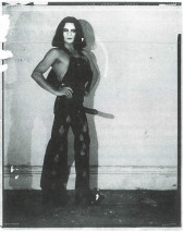 Black and white scan of a photograph featuring a figure, standing upright with their hand on their hips. Their face is starkly painted white with heavy makeup around the eyes and lips. They wear overalls and a strap-on around their hips.