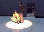 Two figures illuminated by a circular spotlight in an interior space. A mirror leans against the wall directly behind them. One figure stands in a brown suit, their hands behind their back. They are blindfolded. A second, mostly nude figure crouches directly below them.
