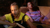 On the right, a woman wearing a purple dress lies down on a couch with her eyes closed. To the left, a man sits on the floor in front of her, wearing a yellow t-shirt and overalls.