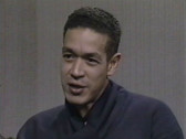 Andres Serrano wearing a dark blue shirt and looking at an off-camera interviewer mid-speech.
