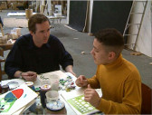 Two people seated at a table covered with assorted art supplies. They talk to each other as they paint or prepare to draw.