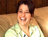 A woman seated on a couch wearing a green button-down shirt captured mid-laughter.